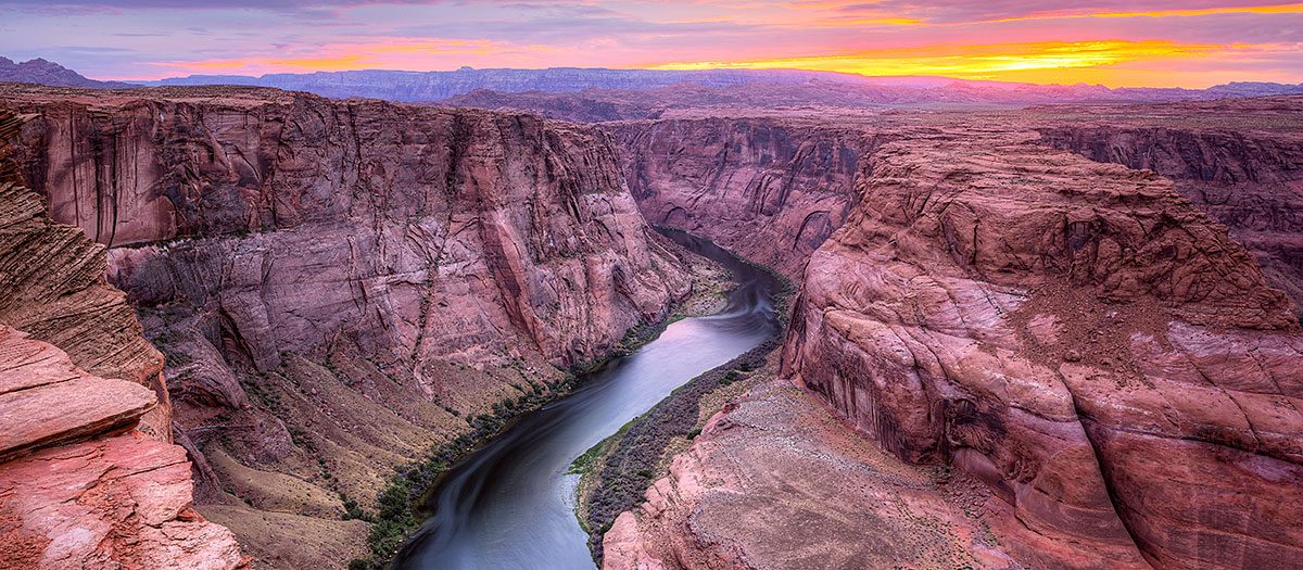 The Colorado River winds its way through red cliffs with a sunset in the background